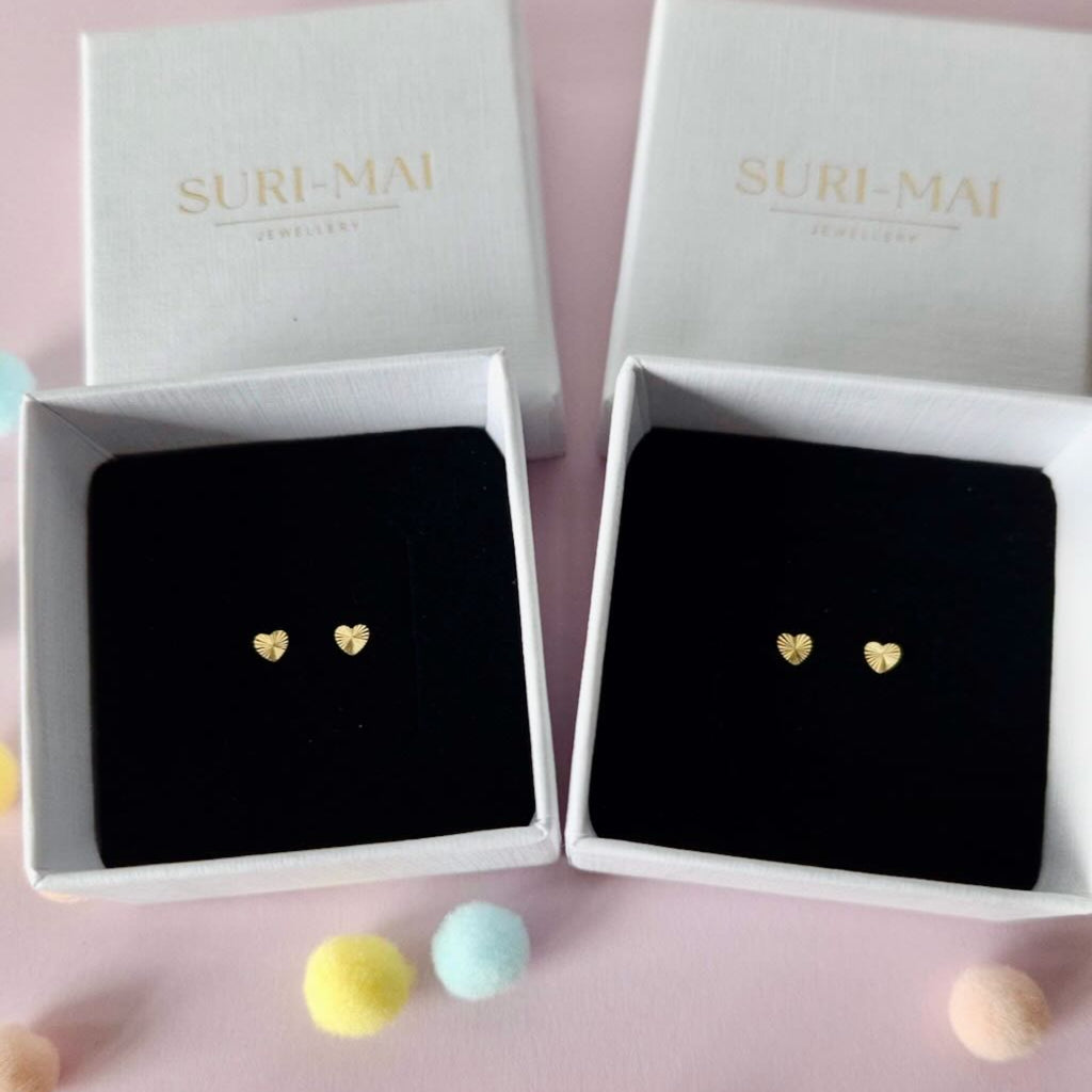 Gold heart-shaped earrings in a 'Suri Mai' box on a pink background with colorful pom-poms.