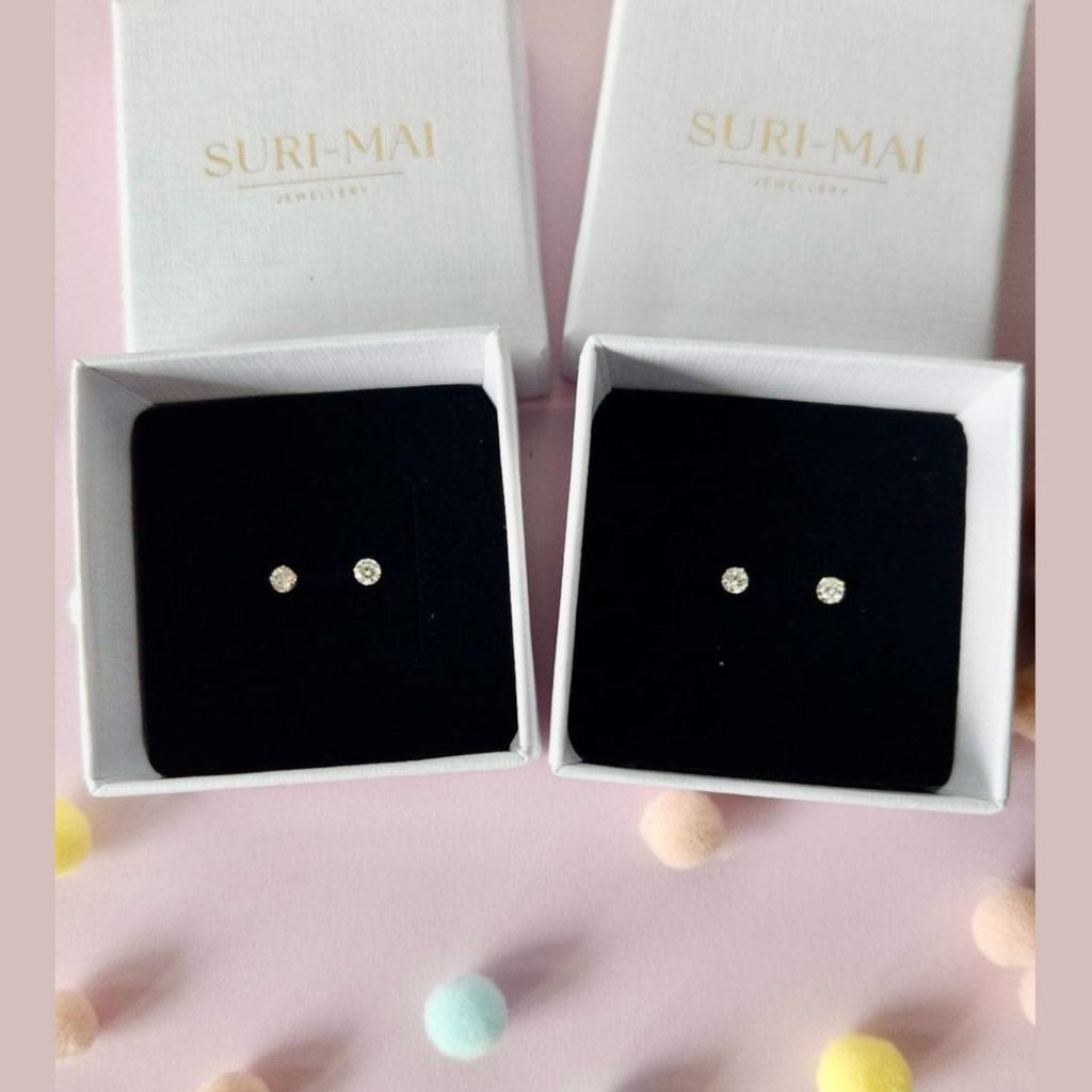 Pair of earrings in a 'SURI-MAI' box on a pink background with colorful balls.