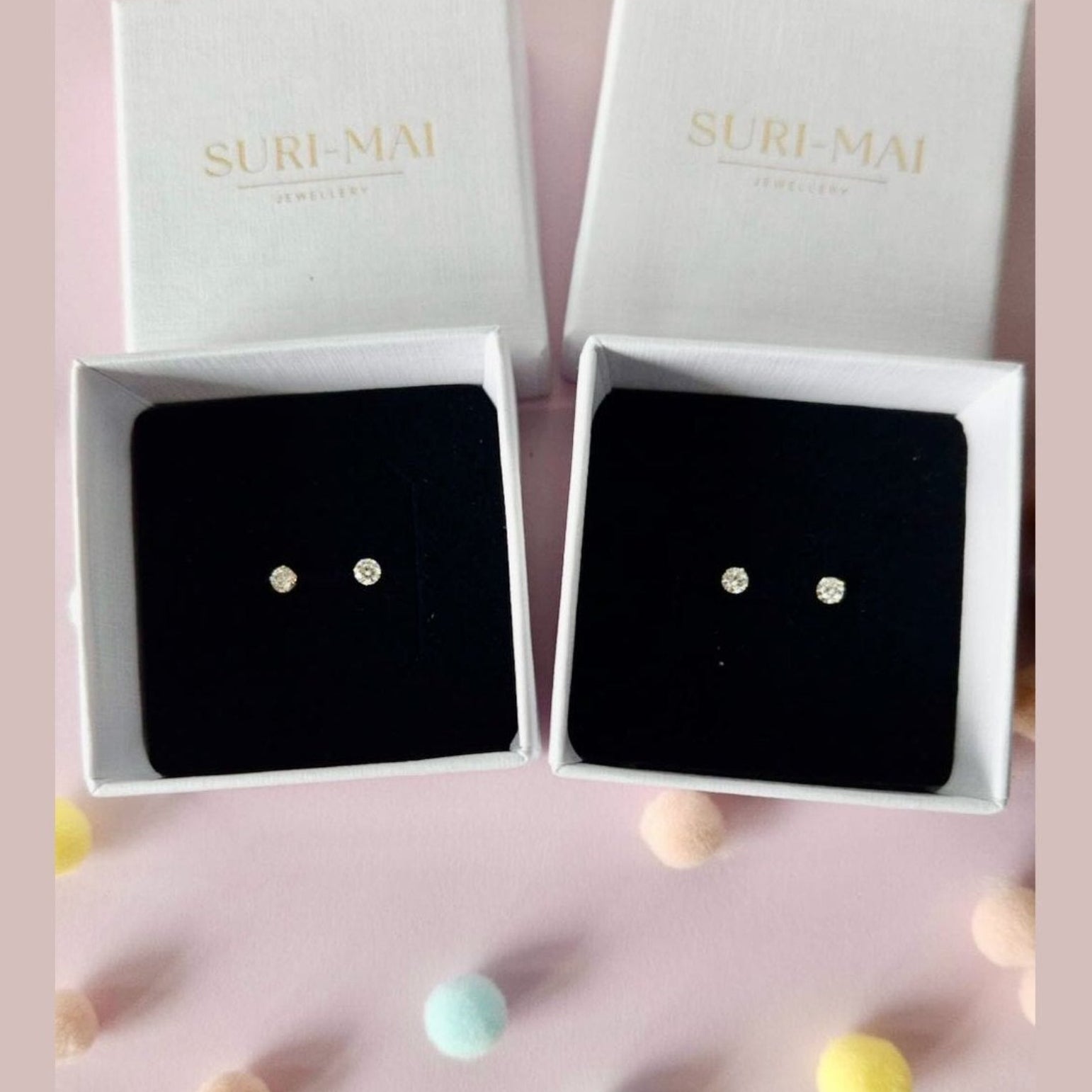 Pair of earrings in a 'SURI-MAI' box on a pink background with colorful balls.