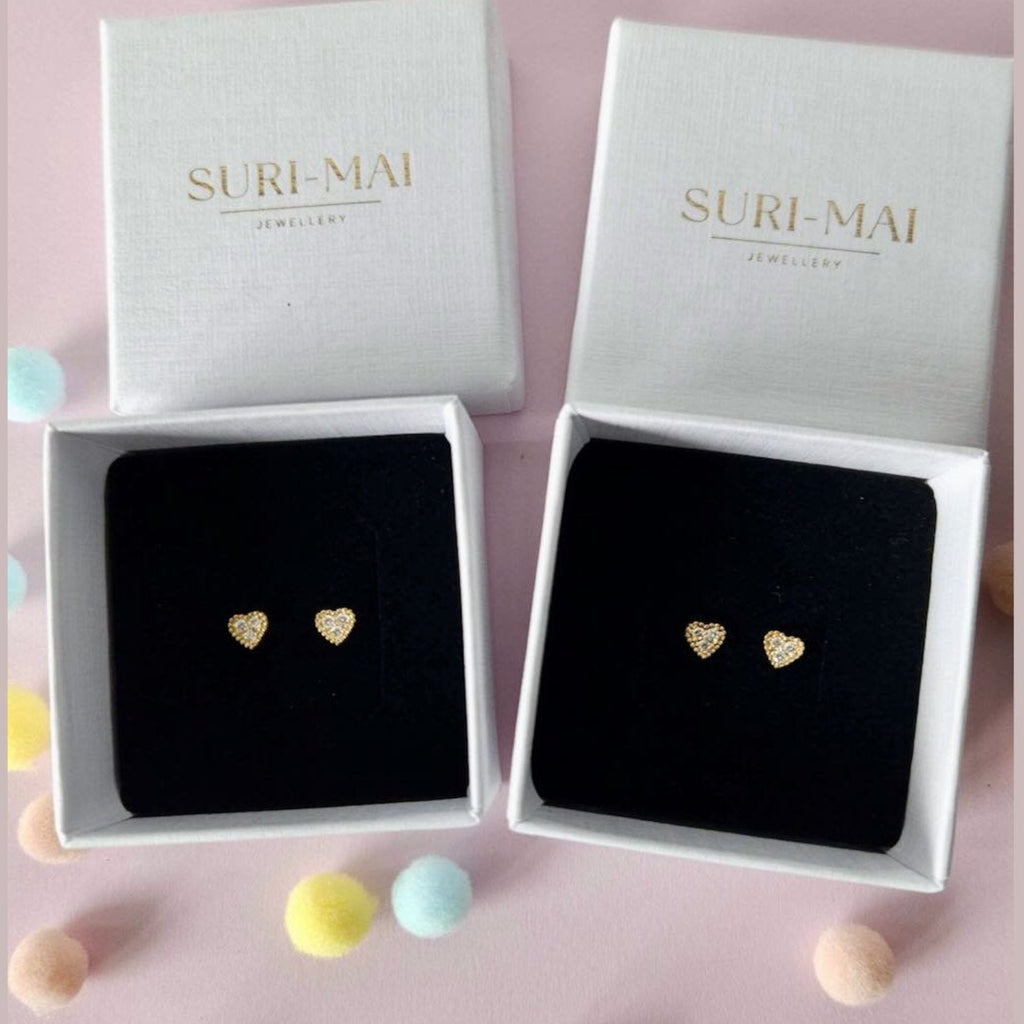 Heart-shaped earrings in Suri-Mai jewelry boxes on a pink background with colorful pom-poms.