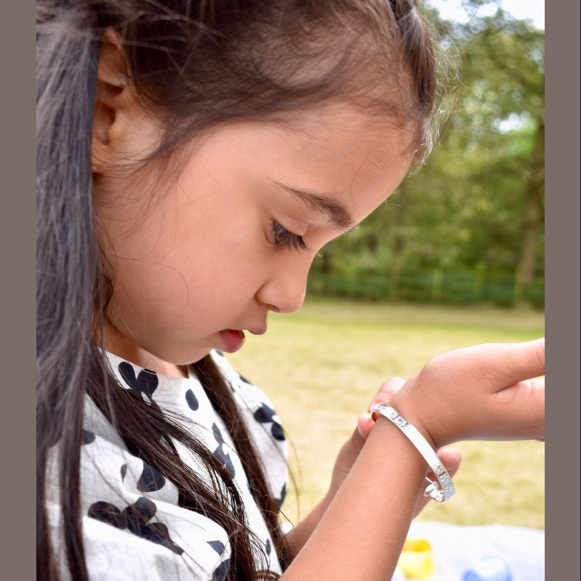 Young girl outdoors wearing a bracelet, with a blurred background of greenery.